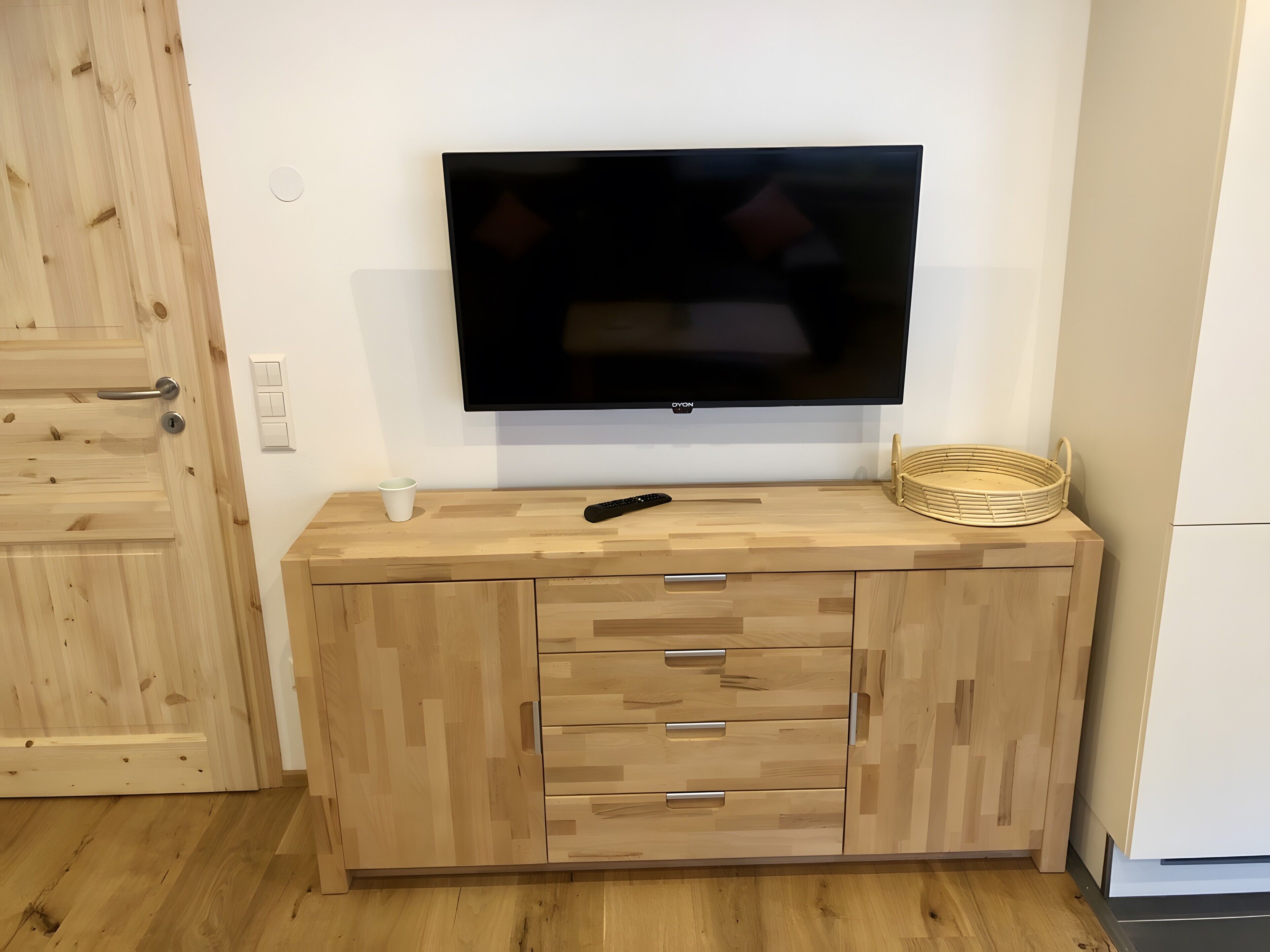 Een houten kast met lades, een televisie erboven bevestigd, een afstandsbediening, een witte beker en een rieten mand.