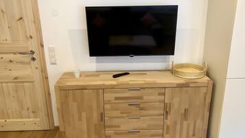 Een houten kast met lades, een televisie erboven bevestigd, een afstandsbediening, een witte beker en een rieten mand.