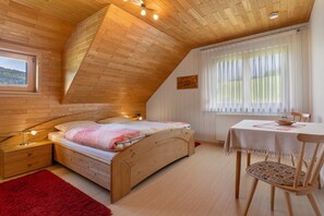 Un dormitorio con techo inclinado, cama, mesita de noche, una mesa con silla y ventana con cortinas.