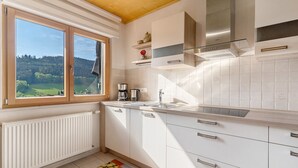 Een keuken met witte kasten, een roestvrijstalen afzuigkap, een raam met uitzicht op een groen landschap en een radiator.