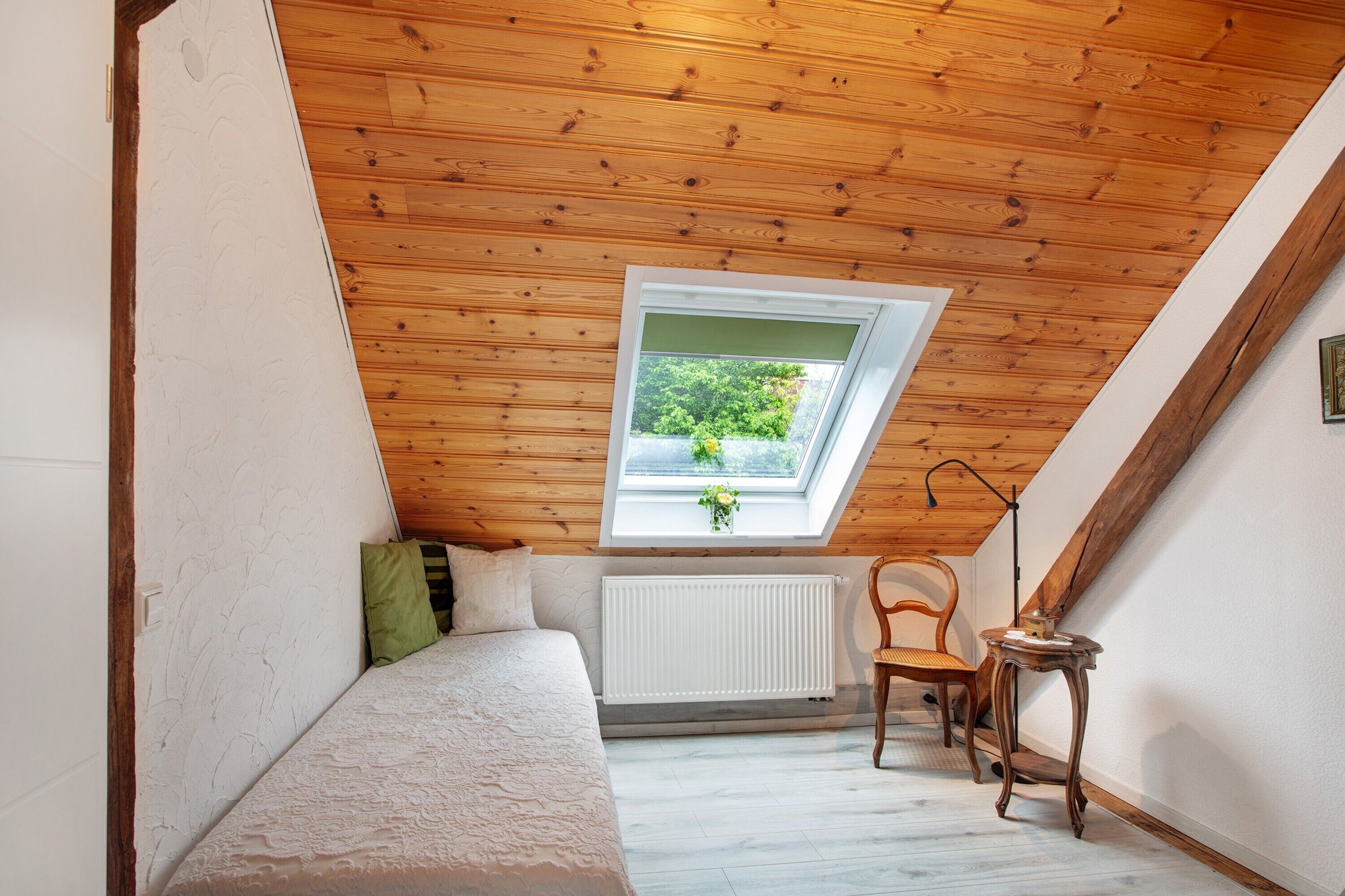 Ein Schlafzimmer mit schrägen Wänden, einem Fenster mit Blick auf Grünflächen, einem Heizkörper, einem Bett und einem Sessel.