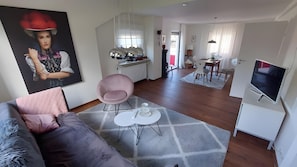 A modern living room with a sofa, a pink chair, a TV, and a dining area with a table and chairs.