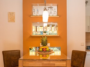 A dining area with a glass table, two chairs, a fruit centerpiece, and two shelves with glassware.