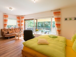 A room with a bed, a sofa, a chair, a window with curtains, and a balcony with plants.