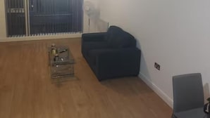 Ein Zimmer mit Holzboden, einem schwarzen Sofa, einem kleinen Tisch mit einer Flasche und einem Fenster mit Jalousien.