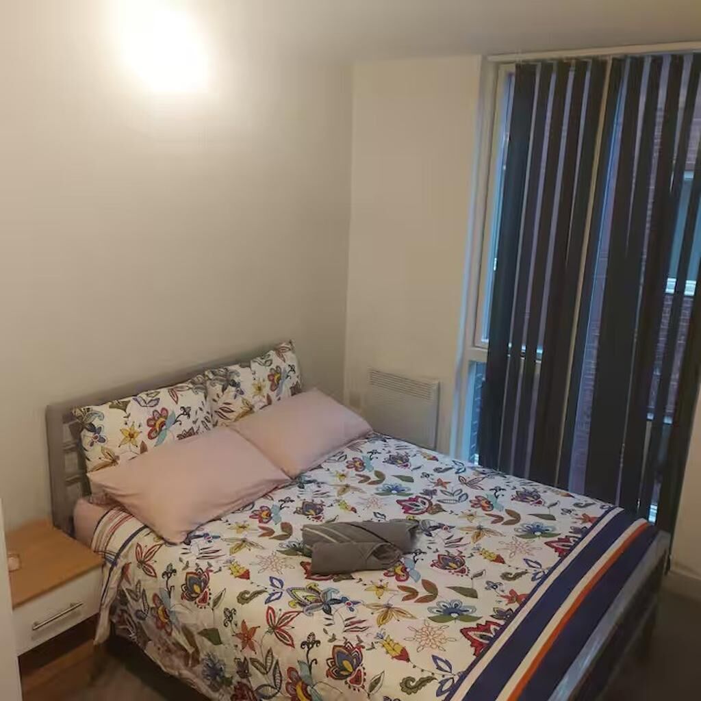 A bedroom with a bed, a nightstand, a window with blinds, and a radiator.