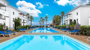A swimming pool with blue water, surrounded by lounge chairs and umbrellas, with white buildings and palm trees in the background. - Appartement Confortable à Côté de la Plage (San-Nicolao)