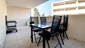Un balcon avec une table en verre et quatre chaises, un petit chariot, et un bâtiment blanc en arrière-plan.