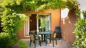 Une terrasse extérieure avec une table et des chaises, entourée de verdure, et une porte vitrée donnant sur une pièce.