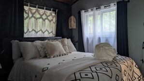 A neatly made bed with patterned bedding and multiple pillows, a decorative hanging light, sheer curtains, and a view of greenery outside. - Charming 2-bedroom cottage in Clarington with hottub, WiFi and AC (Clarington)