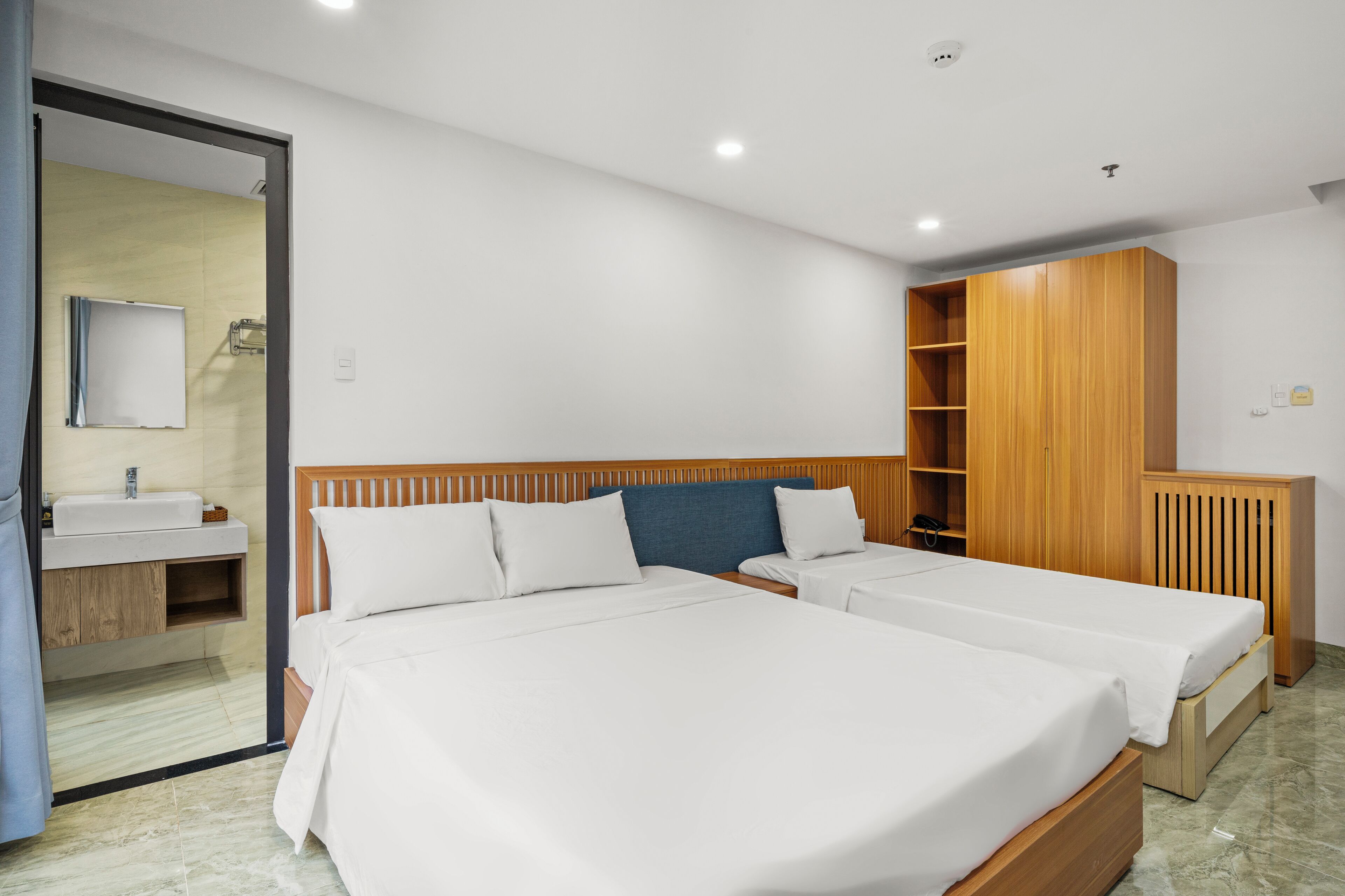 A hotel room with two beds, a wooden wardrobe, a bathroom with a mirror, and a sink.