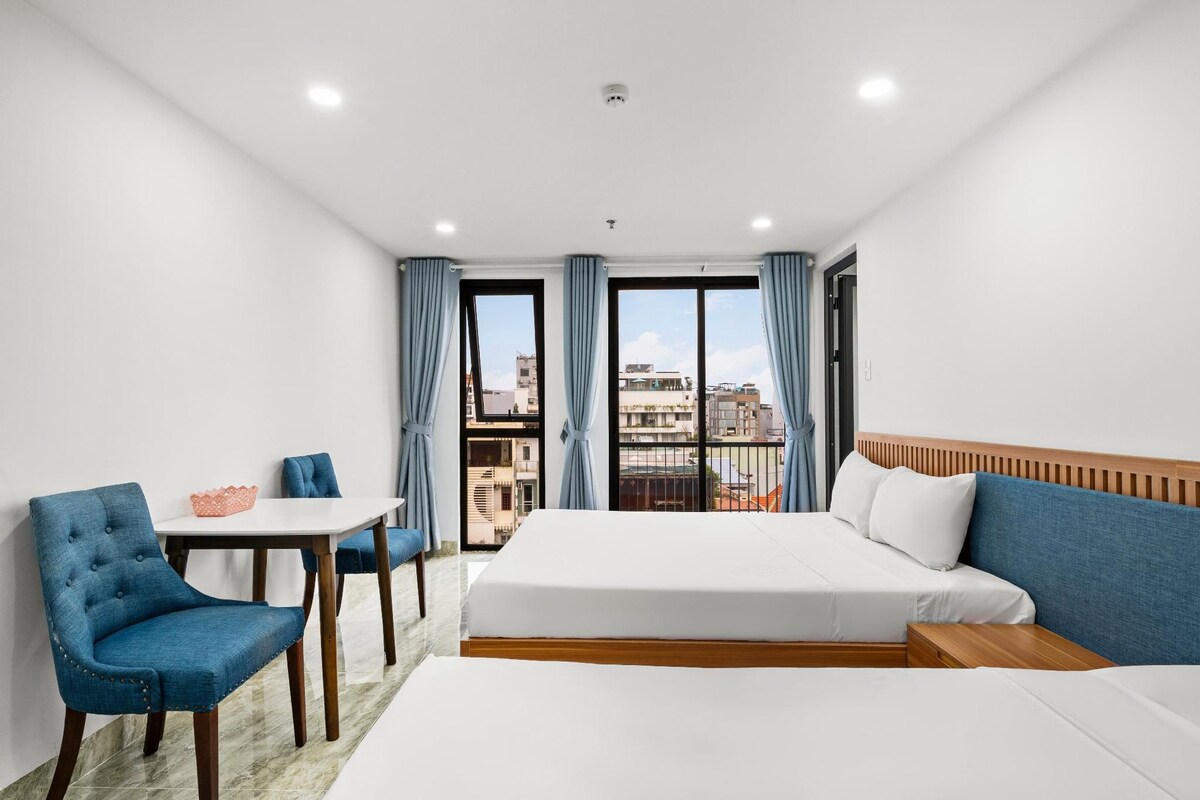 Habitación de hotel con cama, una mesa, dos sillas y vistas a la ciudad a través de amplios ventanales.