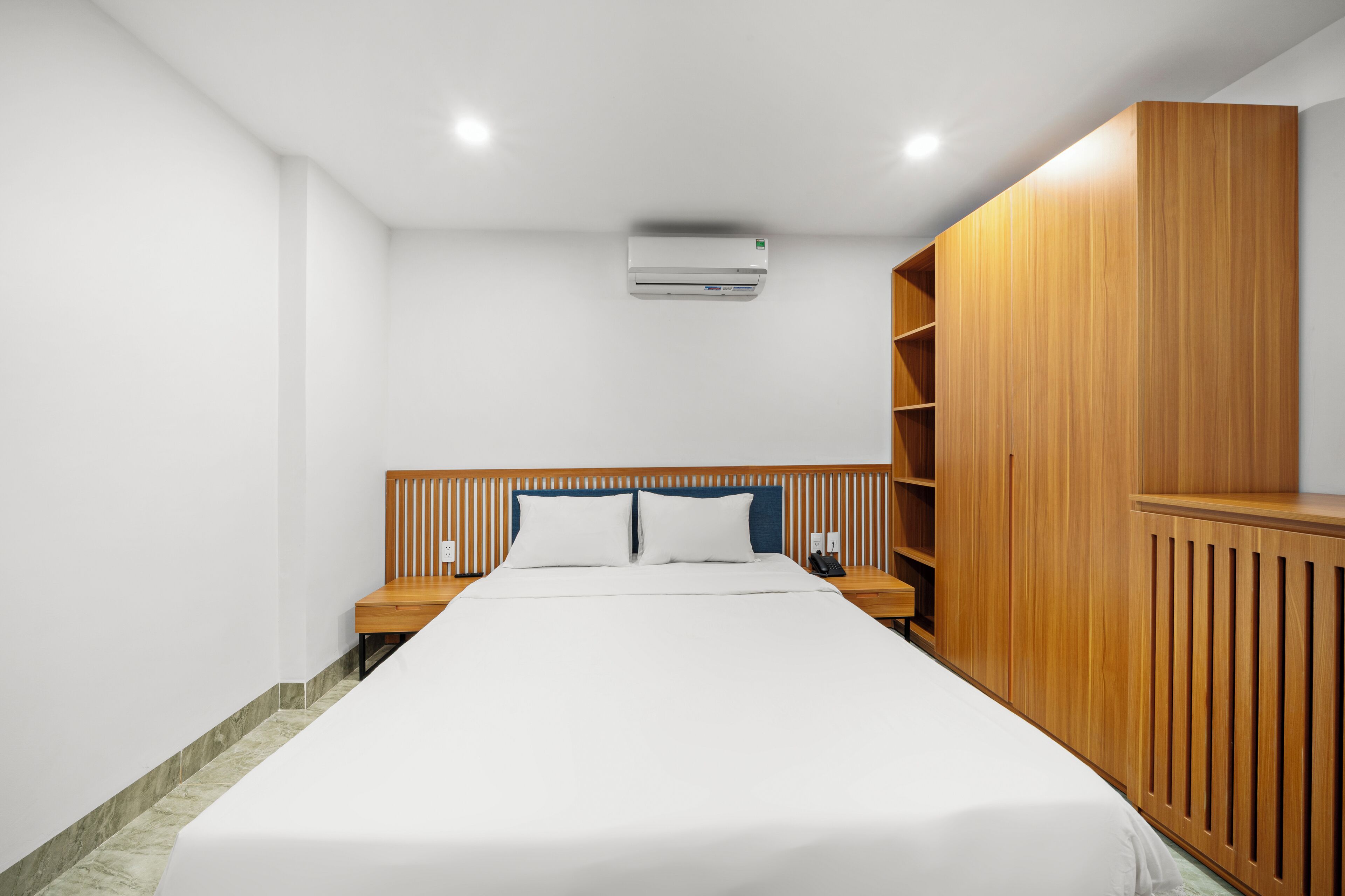 A hotel room with a bed, wooden headboard, a wardrobe, a nightstand, and an air conditioning unit.