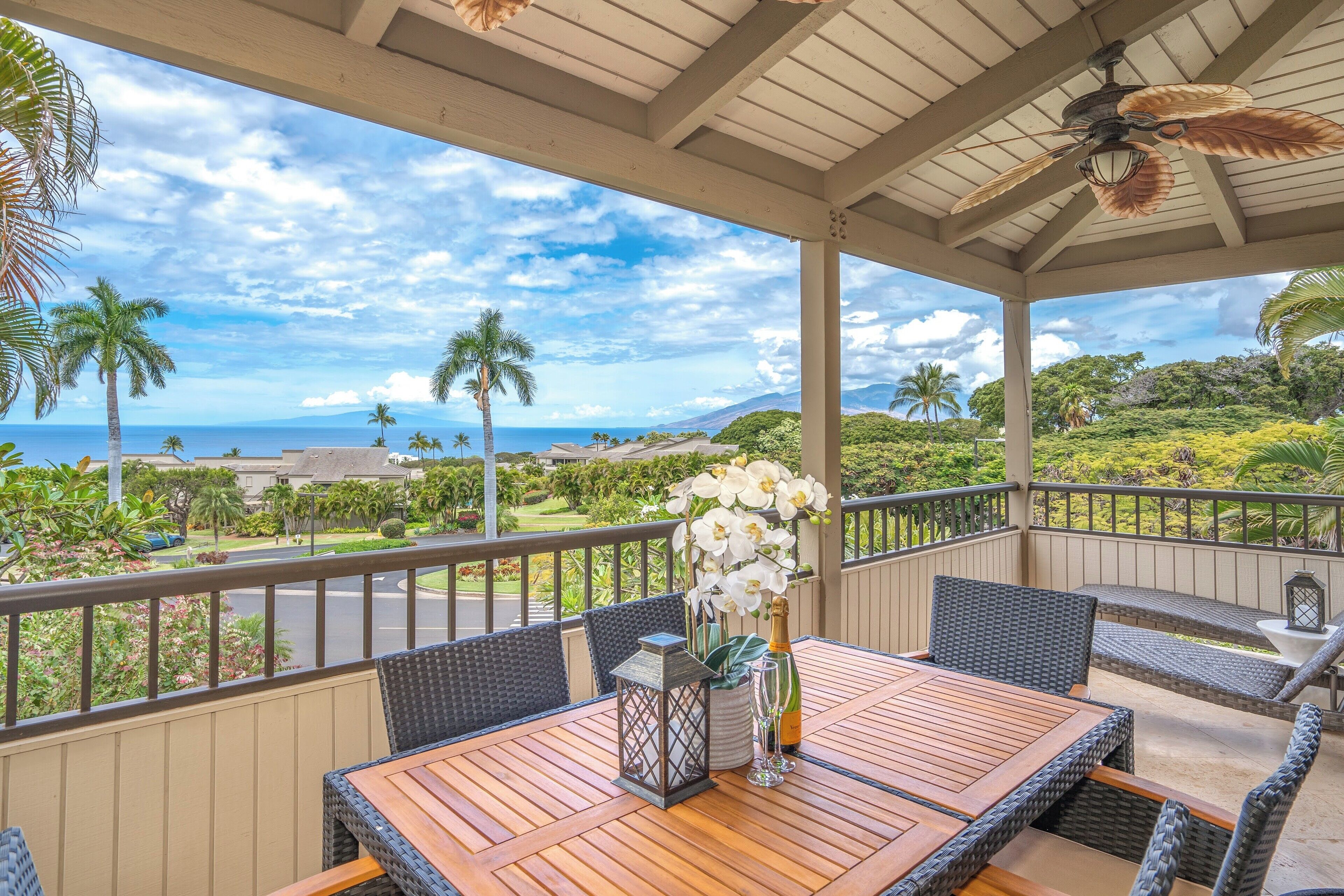 A covered outdoor seating area with wicker furniture, a wooden table, and a view of the ocean and greenery.