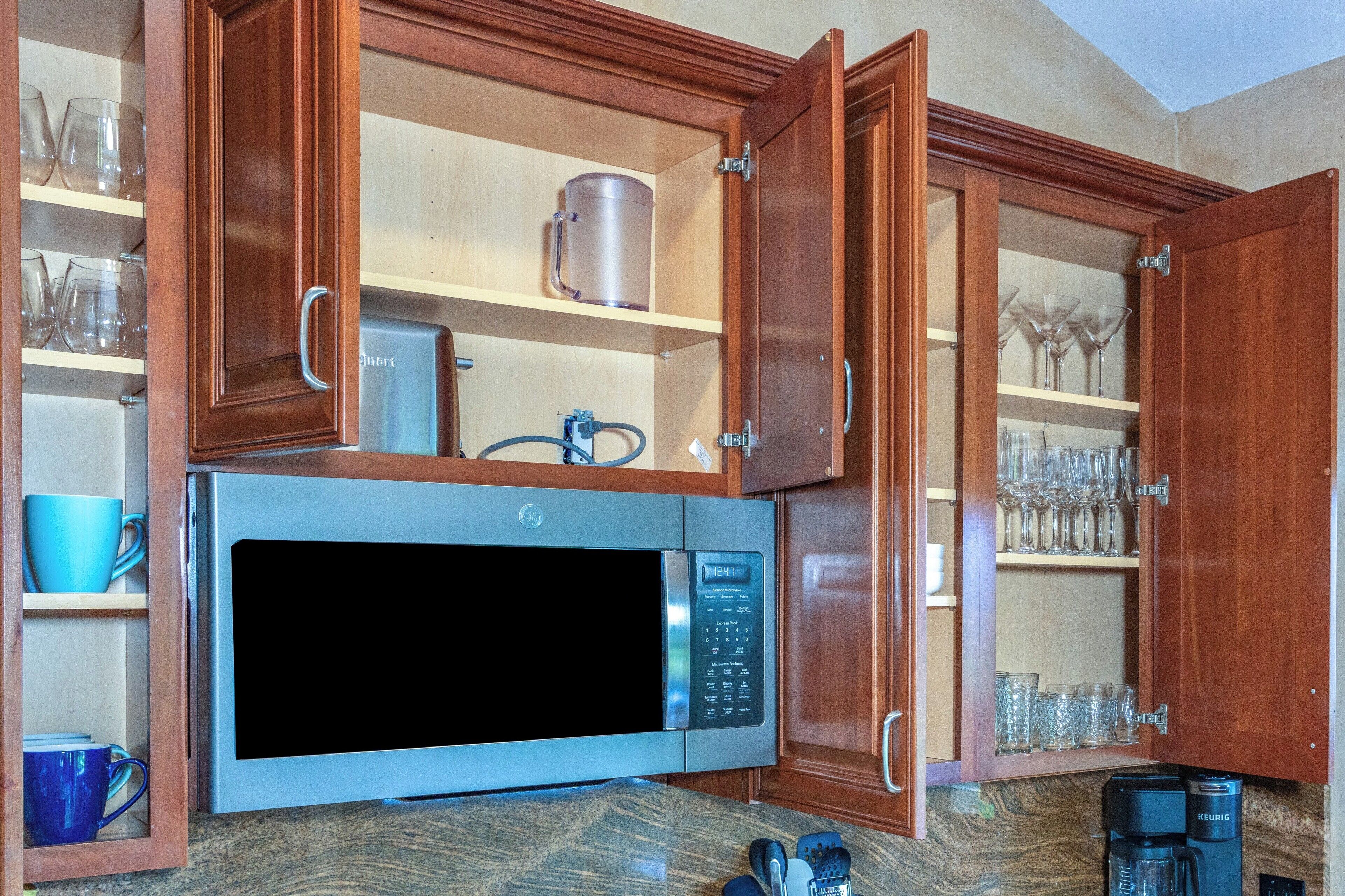 Una cocina con microondas integrado, armarios de madera y estantes con copas y tazas.
