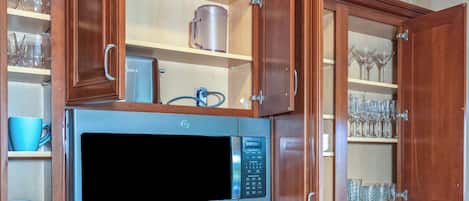 Una cocina con microondas integrado, armarios de madera y estantes con copas y tazas.