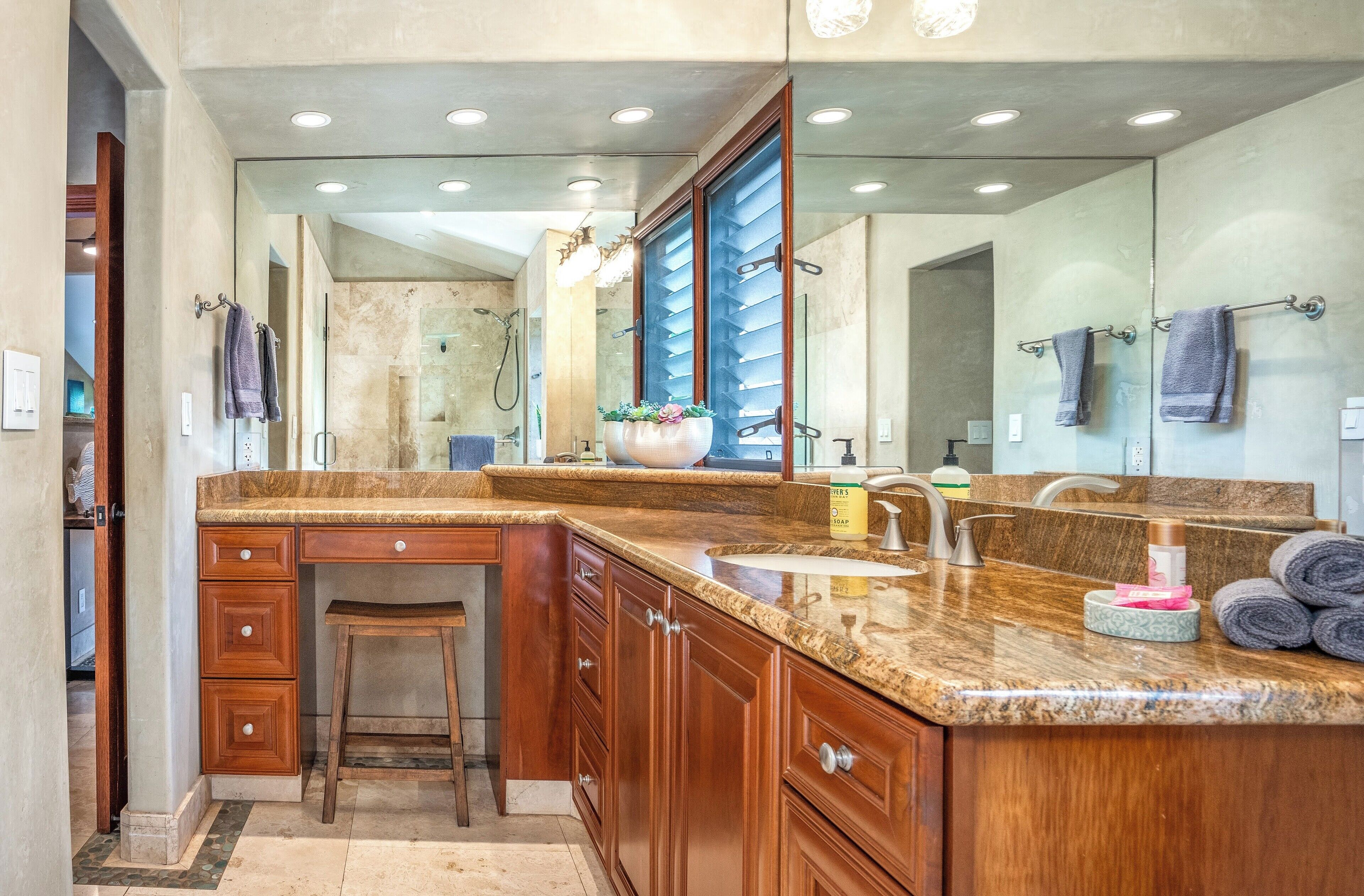 A modern bathroom with a granite countertop, wooden cabinets, a large mirror, and a shower area.