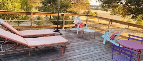 A wooden deck with lounge chairs and a table, surrounded by trees and a field.