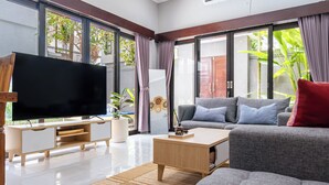 A modern living room with a flat-screen TV, grey sofas, a wooden coffee table, and large windows with curtains.