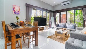 A spacious living room with a dining area, a flat-screen TV, and large windows with curtains.