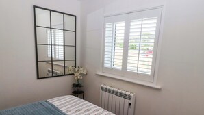 Ein Schlafzimmer mit einem Bett, einem Heizkörper, einem Fenster mit Jalousien, einem Spiegel und einer Zimmerpflanze.