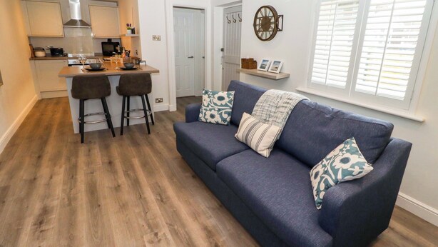 Ein modernes Wohnzimmer mit einem dunkelblauen Sofa, zwei passenden Sesseln und einem Couchtisch. Im Hintergrund ist eine Küche mit weißen Schränken und einer Edelstahl-Dunstabzugshaube zu sehen.