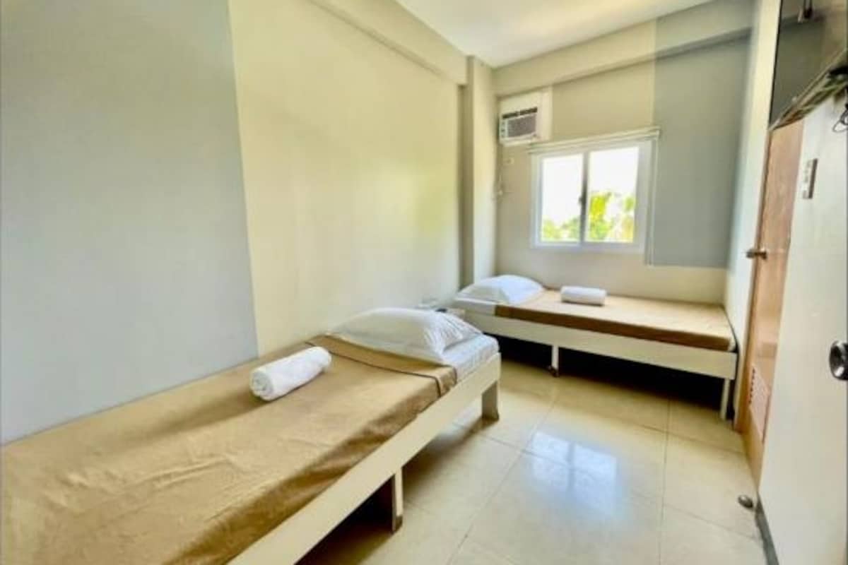 A small, single-room hotel room with two single beds, a window, and an air conditioning unit.