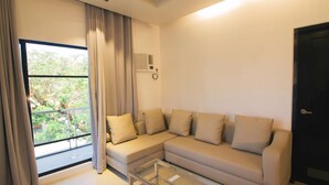 Un salón con un sofá beige, una mesa de centro de vidrio y una ventana con cortinas.