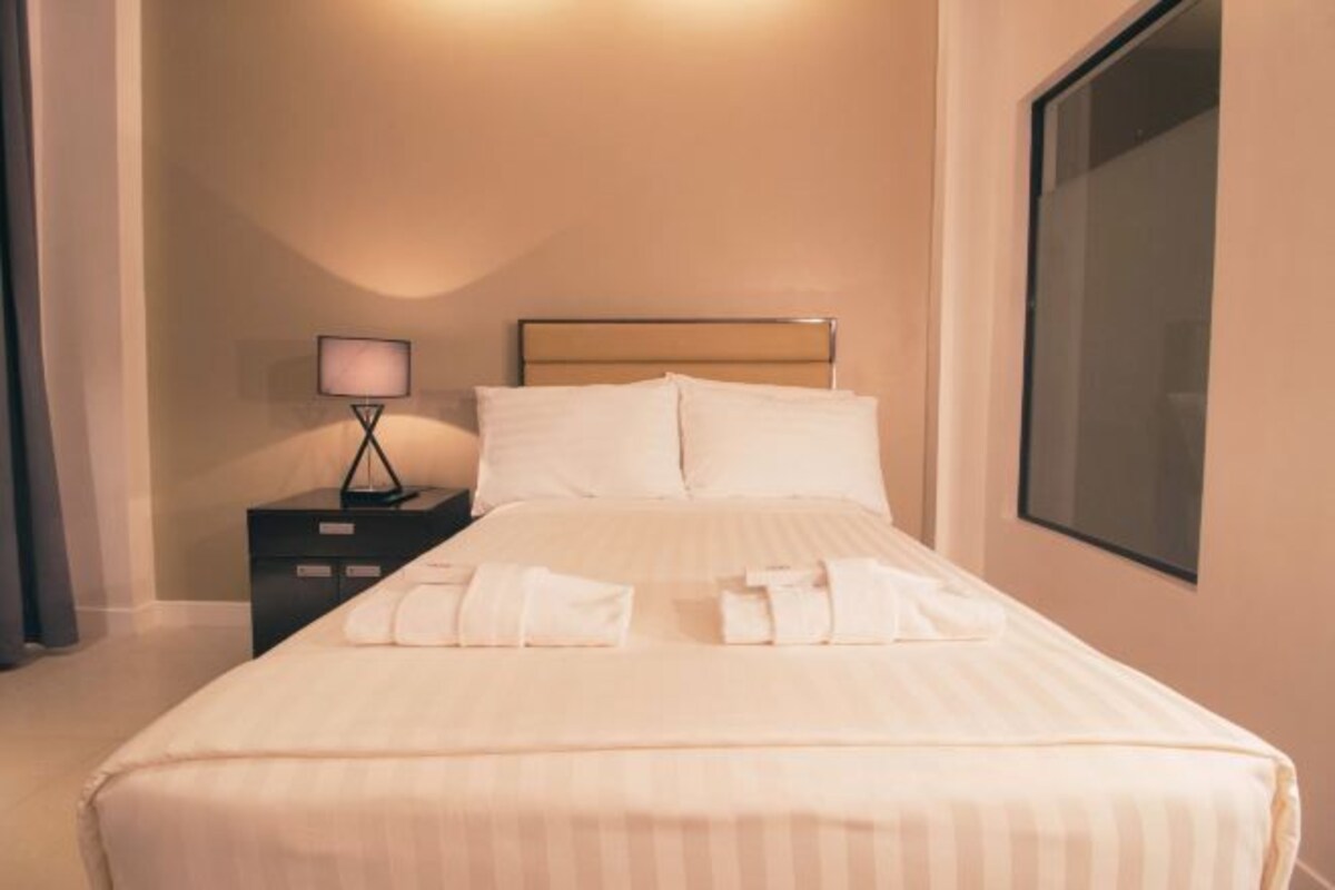 Una cama bien hecha con sábanas y toallas blancas, una lámpara de noche y un ventanal amplio.