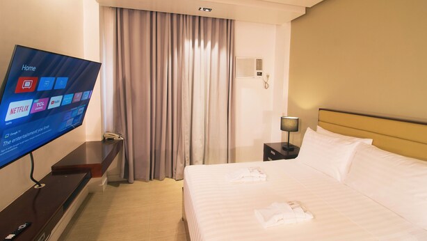 Habitación de hotel con cama, televisión mostrando la pantalla de inicio, mesita de noche, lámpara y ventana con cortinas.