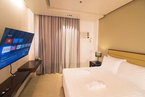 A hotel room with a bed, a TV displaying the home screen, a bedside table, a lamp, and a window with curtains.