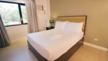 A neatly made bed with white linens and towels, a bedside table with a lamp, and a window with curtains.
