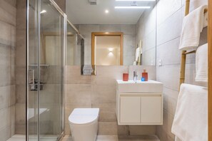 Une salle de bain moderne avec une douche à parois de verre, des toilettes blanches, un lavabo blanc avec un miroir au-dessus, et un porte-serviettes en bois.
