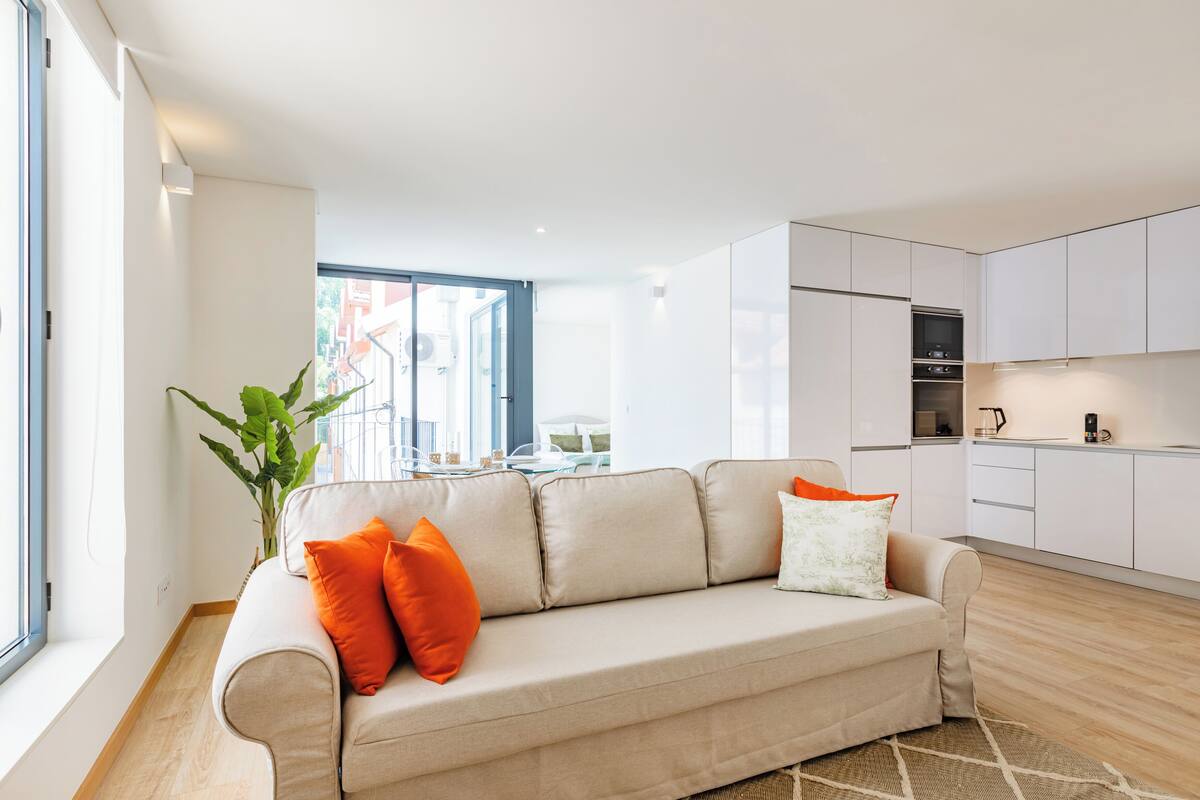 Ein modernes Wohnzimmer mit einem beigefarbenen Sofa, orangefarbenen Kissen, einem großen Fenster und Ausblick nach draußen.