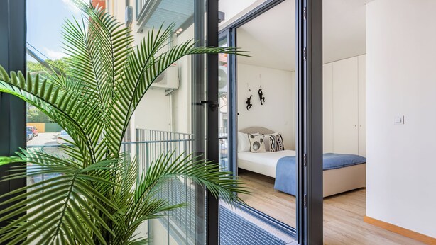 A modern apartment with a sliding glass door leading to a balcony with a potted plant. Inside, there is a bed with a blue mattress and a white wall with a few decorative items. - Duque de Saldanha Residences (Porto)
