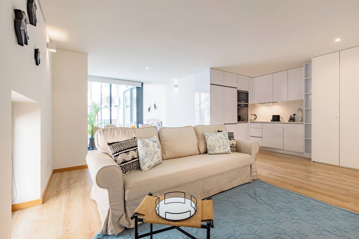 Ein modernes Wohnzimmer mit einem beigefarbenen Sofa, einem kleinen Couchtisch und einer Küchenzeile mit weißen Schränken und einer Spüle.