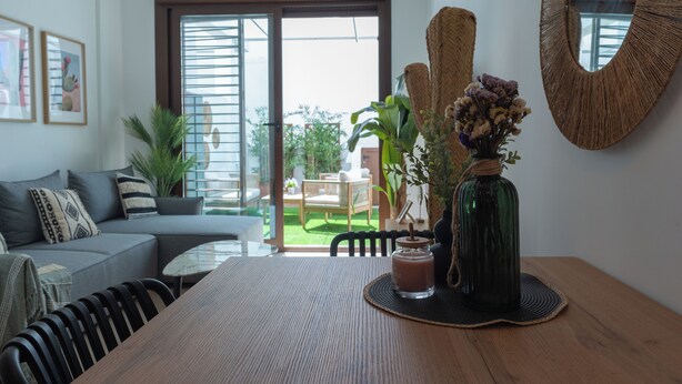 A modern living room with a sofa, a dining table, and a large mirror. There is a view of a garden through the glass door.