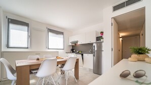 Una cocina moderna con muebles blancos, un refrigerador de acero inoxidable y una zona de comedor con una mesa de madera y sillas blancas.