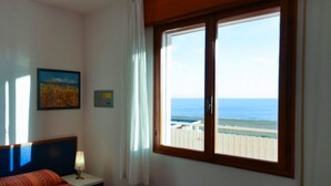 Une chambre à coucher avec un lit, une table de chevet, une lampe, un tableau et une fenêtre donnant sur la mer et une clôture blanche.