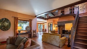 A spacious living area with a sofa, a coffee table, and a fireplace. There is a staircase with ornate railings leading to an upper level. - My Rental Homes - Villa Brin with tennis court, private sandy beach and dock (Lesa)