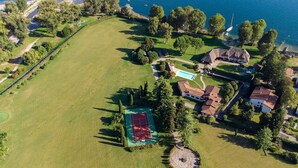 An aerial view of a property with a large grassy field, a tennis court, a swimming pool, and several buildings including a house with a thatched roof and a white building with a red roof. - My Rental Homes - Villa Brin with tennis court, private sandy beach and dock (Lesa)