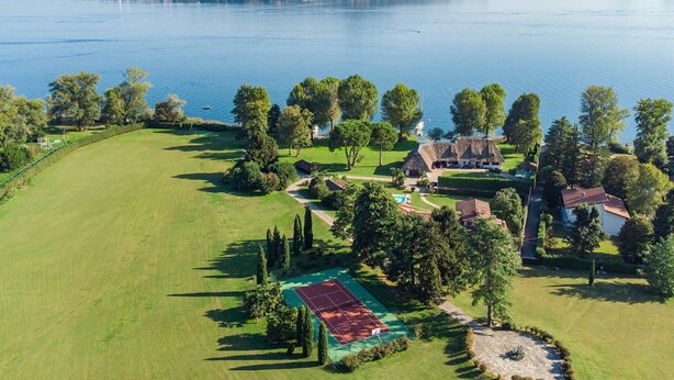 An aerial view of a lakeside property with a tennis court, a thatched-roof house, and a large green lawn. - My Rental Homes - Villa Brin with tennis court, private sandy beach and dock (Lesa)