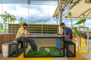 Two men sitting at a table with a soccer ball on a green artificial turf, surrounded by potted plants and a yellow railing.