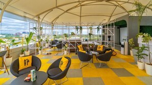 Un espace de détente moderne avec un plafond haut, un sol à carreaux jaunes et gris, des chaises noires avec des coussins jaunes et des plantes en pot.