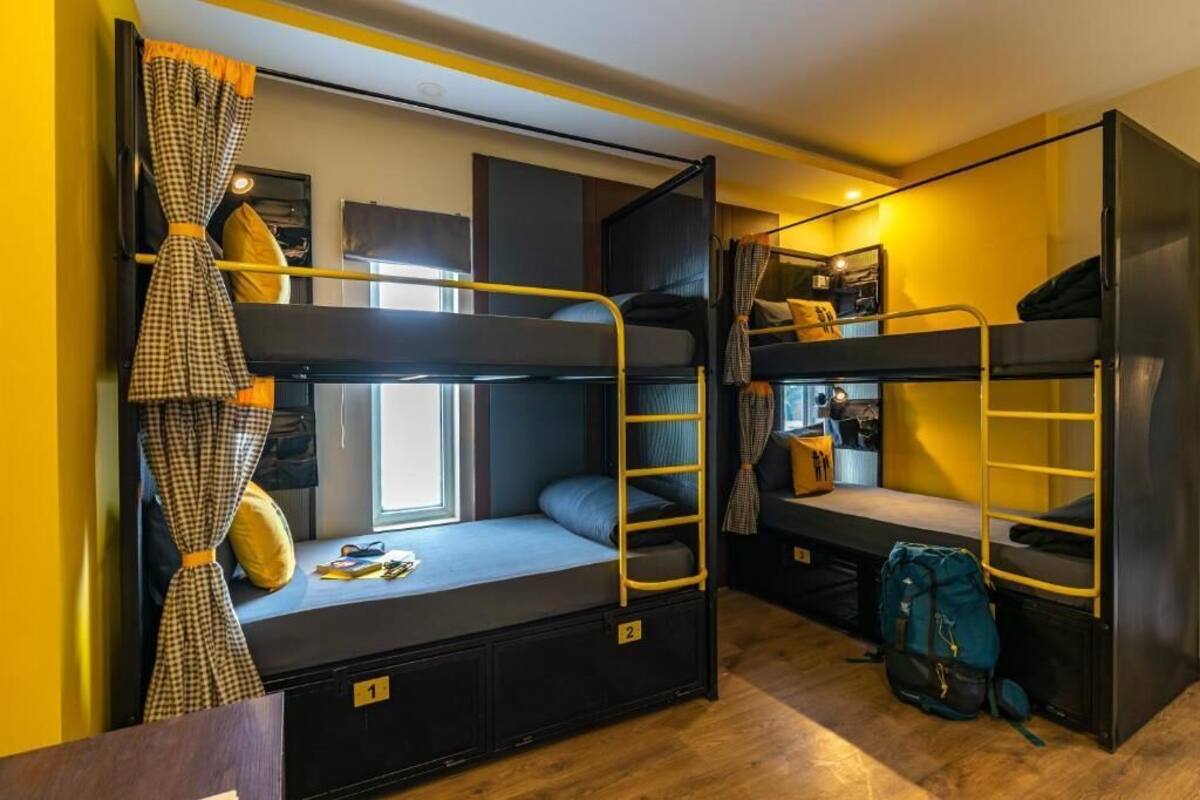 Ein Schlafsaalzimmer mit Etagenbetten, einem Fenster mit Vorhängen, einem Schreibtisch und einem Rucksack auf dem Boden.