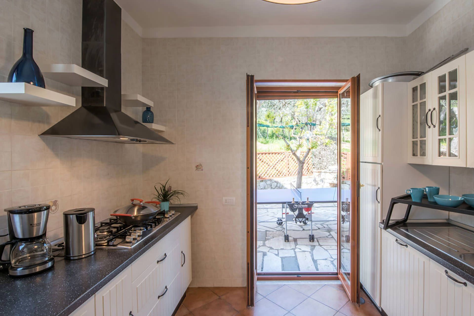 Una cocina con armarios blancos, una estufa a gas, una cafetera y vistas a un patio con mesa y sillas.