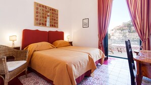 A hotel room with a bed, a wicker chair, a table, and a view of buildings through the window. - My Rental Homes - Casa Li Galli  5mins' walk from Positano center, terrace, sea view (Positano)