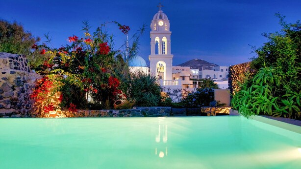 A pool with a view of a white building with a clock tower, surrounded by colorful plants and a stone wall. - Mansion Kyani with pool, large terrace and sea view (Fira)
