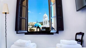 A hotel room with an open window revealing a view of a white building with a clock tower and a blue dome. There are two white towels on the bed. - Mansion Kyani with pool, large terrace and sea view (Fira)