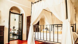 Ein Bett mit Baldachin und hölzernem Gestell, dazu weiße Bettwäsche, ein Holfschrank und ein Spiegel in einem Zimmer mit rotem Boden und einer Tür zu einem weiteren Raum.